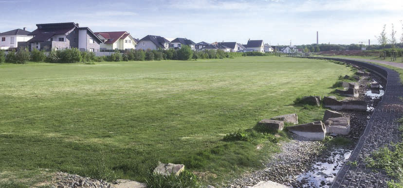 Kommune setzt auf Versorgung mit Wasser-Wasser-Wärmepumpen Foto des Wohngeboets Eichholz: in der Ferne sind Häuser zu sehen, davor grüne Wiese und der Bachlauf.