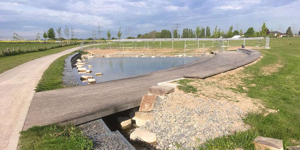 Sicht auf einen künstlichen Bachlauf und Teich als Teil eines Nahwärmenetzes, mit Holzsteg und umliegender Grünfläche.